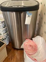 Front view of a stainless steel trash can with a black automatic sensor lid and rolls of pink-tinted and white trash liners placed at the base.