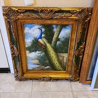 Full view of peacock painting with ornate gold frame leaning against wall.