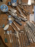 Overview photo depicting an assortment of silver-plated flatware and specialized serving utensils on a wooden table.