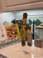 Photo showing a section of the spice rack with three decorative glass bottles filled with spices or grains, and a glass jar containing almonds, accompanied by a cookbook in the background held by a decorative metal stand.