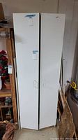 Front view of tall white melamine storage cabinet with two doors, showing both door handles and some marks on the surface.
