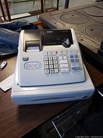 Front angled view of Casio PCR-T275 cash register showing keypad, receipt printer, and cash drawer on wooden surface near wood stove.