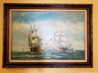 Full front view of the framed naval ships battle painting hanging on a wall, showing the two large sailing warships on ocean with cloudy sky.