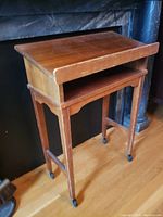 Front left angled view showing slant-top surface, open storage shelf, and four legs with caster wheels on wooden floor.