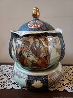 Front view of the lidded porcelain biscuit jar decorated with detailed Japanese Satsuma style artwork featuring multiple traditional figures and intricate patterns in dark and gold tones. The jar has a uniquely shaped body, a matching lid with a bird-shaped golden knob, and a decorative base.