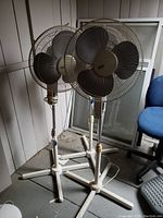Three beige standing fans with metal cage guards and tripod base in a corner setting showing overall condition and setup.