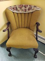 Front view of vintage armchair showing mustard-yellow textured upholstery, carved wooden arms with wear, claw feet, and floral fabric cover on top back.