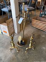 Brass fireplace utensil set including stand with brush, shovel, and poker, plus two fire dogs on the floor.
