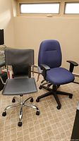 Photo showing both office chairs in a room with a window and carpeted floor. The blue fabric swivel chair is on the right, and the black faux leather chair with metal frame is on the left.