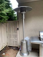 Full view of Sunverse freestanding patio heater showing stainless steel perforated body, base, pole, and circular reflector top. Positioned outdoors near a grill and wooden fencing.