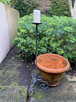View of terra cotta planter next to tall black metal candle holder with white candle outdoors near bushes