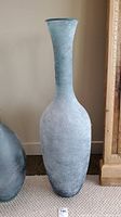 Full front view of tall narrow-necked glass vase with frosted blue-grey matte finish sitting on floor beside a wooden frame.