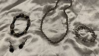 Photo of all four costume jewelry pieces on white fabric: a twisted silver-tone chain necklace, a silver-tone linked bracelet, a black and white beaded bracelet, and matching dangle earrings.