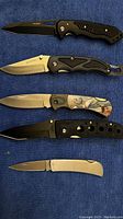 Five folding knives laid flat on a blue cloth showing blades open for viewing design and condition.