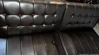 Close-up front view of black leather-look vinyl futon showing tufted backrest and seat details.