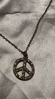 Close-up photo of a silver-tone peace sign pendant necklace embellished with small multicolored rhinestones lying on a satin-like fabric.