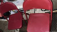 Two unfolded red steel folding chairs with padded seats and backs. Shows the frame design, red color, and condition of the padding and metal parts.