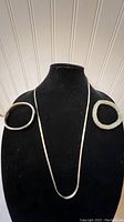 Front view showing the sterling silver chain necklace centered on a black display bust with two bracelets on shoulders; left bracelet is solid metal, right bracelet is white beaded.