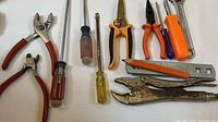 Wide view of the assortment of tools including pliers, vice grips, screwdrivers, utility knife and metal file arranged on white surface.
