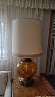 Full view of large amber glass table lamp with cream fabric shade, situated beside a couch and lace curtains in the background.