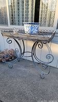 Distressed metal garden stand placed outdoors against a window with lace curtains, showing full view of stand including tray top and scroll legs.