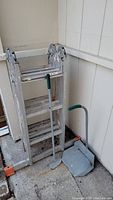 Photo of aluminum extension ladder folded up and standing against a wall with a metal garden scoop in front.