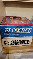 Two stacked original Flowbee boxes, one blue and one white.