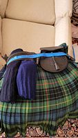 Photo showing folded plaid kilt in green tartan with a pair of knitted dark socks and a black leather sporran with tassel resting on top.