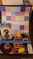 Overview of the lot showing compass in packaging, Ryobi charger, screwdriver, tape measures, electrical tape, and folding ruler in a box with patchwork patterned background.