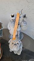 Two outdoor figurines on a glass table against textured wall: one of a standing grizzly bear holding a branch with an acorn ornament, the other a seated bulldog figurine, both showing some wear