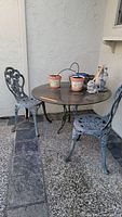 Overview of the round metal outdoor table and two matching chairs placed on a stone patio floor against a stucco wall. The table holds several ceramic flower pots and decorative items.