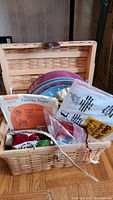Open wicker sewing basket with lid open displaying assorted sewing supplies including pin cushions, tracing paper packets, and various bags of supplies.