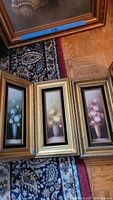 Photo showing all three framed flower paintings arranged on a rug and hardwood floor. The paintings feature blue, yellow, and pink floral arrangements in tall narrow gold frames.