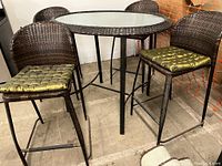 Bar height outdoor patio dining set with a round frosted glass top table and four dark brown braided wicker chairs. Green silk cushions on chairs visible. Rust on metal legs evident.