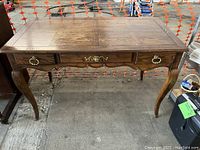 Front view of wooden replica Queen Anne style desk with three front drawers and curved legs on concrete floor.