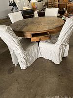 Full view of round solid oak dining table with four high back chairs covered in white damask slipcovers, one drawer open showing storage.