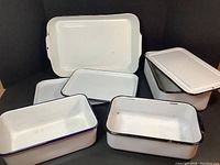 Photo showing two large rectangular white enamel trays with black trim stacked and three deep enamel pans in front with black and blue rims.