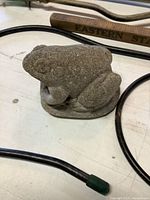 Small concrete frog garden decoration resting on a surface with a wooden handle and black wire frames visible