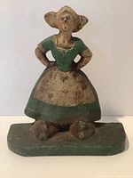 Front view showing the painted lady in a green dress and bonnet, hands on hips, standing on a base.