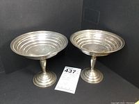 Pair of sterling silver footed bon bon dishes with thumb-sized wide circular bowls, shown from front angle on black background.