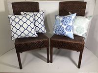 Two brown rattan dining chairs with four assorted decorative pillows placed on them.