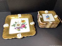 Square decorative plates with scalloped edges, heavy gold border and white raised accents, each featuring colorful flower paintings in center.