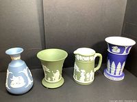 Four Wedgwood Jasperware pieces shown together: blue bud vase, green cup, small green pitcher, small blue vase, each with white classical relief decoration.