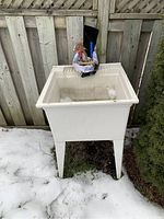 Front view of basement sink placed outdoors on snow-covered ground by wooden fence. Shows sink, faucet with handles, and plumbing parts inside basin.