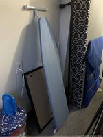 Ironing board with blue cover leaning against the wall, showing the full length and support stand.