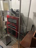 Photo showing two glass display cabinets positioned side by side. Cabinets have metal frame construction with clear glass panels and several internal glass shelves. Red tool chest and other items visible behind the cabinets.