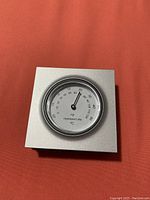 Photo of a small silver square thermostat with a round analog temperature dial in Celsius, placed against a reddish background.