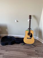 Full view of El Dégas acoustic guitar on stand beside the soft case on the floor against a beige wall.