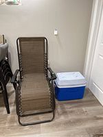 Gravity reclining patio chair and blue and white cooler placed on wood floor indoors.