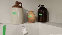 Three jugs on a shelf: large ceramic two-tone jug, smaller pottery jug, dark brown glass jug with handle and nozzle. Green stickers on each showing lot numbers. Tag attached to shelf.